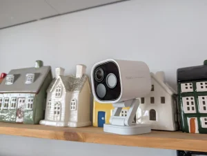 Biała inteligentna kamera Aqara na drewnianej półce pośród dekoracyjnych ceramicznych domków.