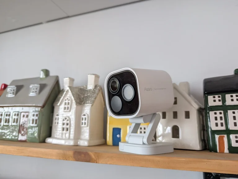 Biała inteligentna kamera Aqara na drewnianej półce pośród dekoracyjnych ceramicznych domków.